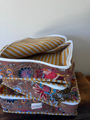 A stack of folded patterned quilts or blankets with white piping, including a mustard and white striped design and a brown floral patchwork design, photographed indoors against a plain wall and wooden surface.