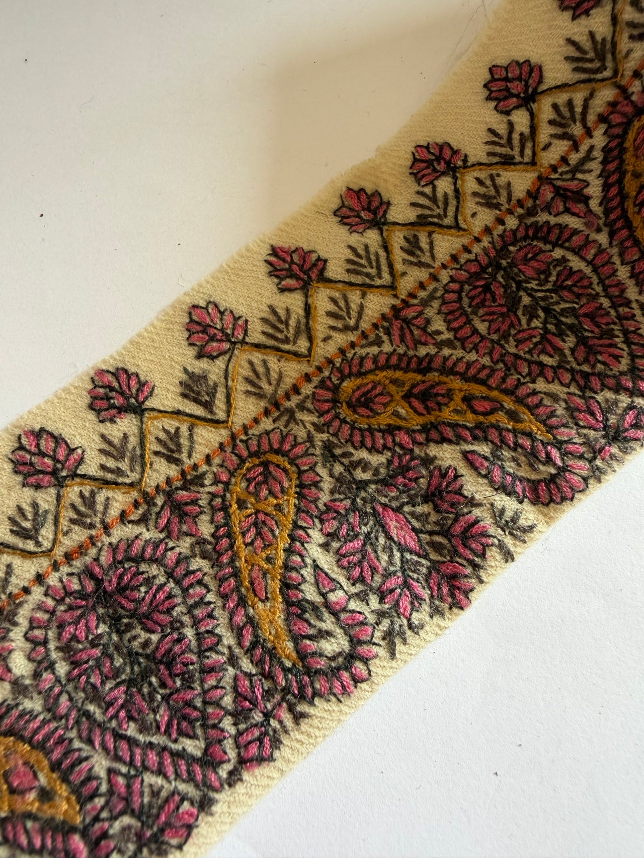 A close-up of a woven fabric trim featuring a classic paisley pattern in dusty pink, mustard yellow, and black on a cream background.