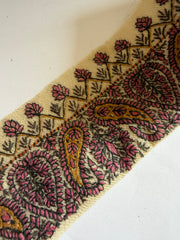 A close-up of a woven fabric trim featuring a classic paisley pattern in dusty pink, mustard yellow, and black on a cream background.