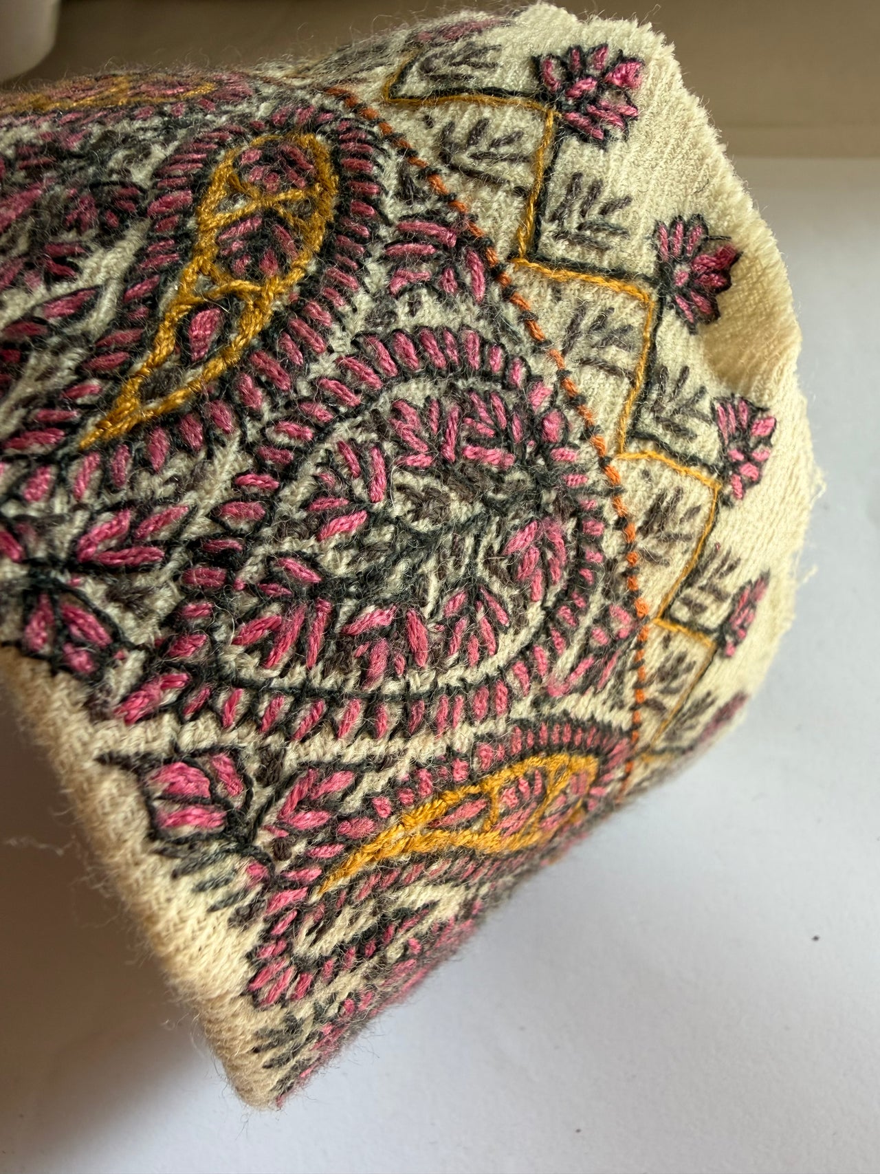 A close-up of a cream-colored woven textile, likely a traditional embroidered cap or hat, featuring intricate paisley and floral patterns in pink, olive green, and mustard yellow.