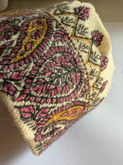 A close-up of a cream-colored woven textile, likely a traditional embroidered cap or hat, featuring intricate paisley and floral patterns in pink, olive green, and mustard yellow.