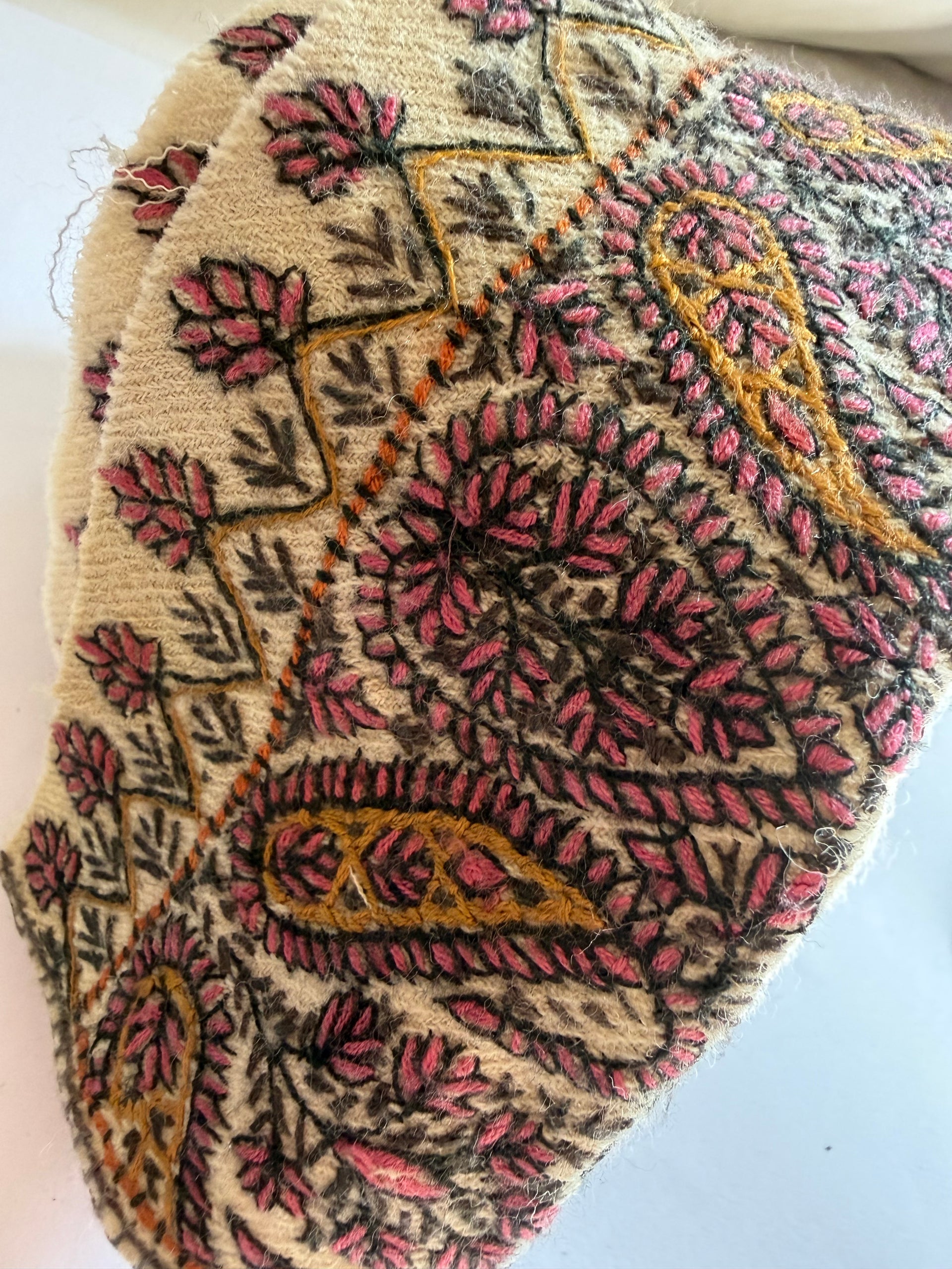 A close-up of a textured fabric or rug featuring a detailed paisley and floral pattern in pink, gold, and dark brown on a cream background.