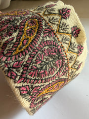 A close-up of a knitted or crocheted textile piece, likely a hat or bag, featuring a cream base with pink, black, and mustard yellow embroidered paisley and floral patterns.
