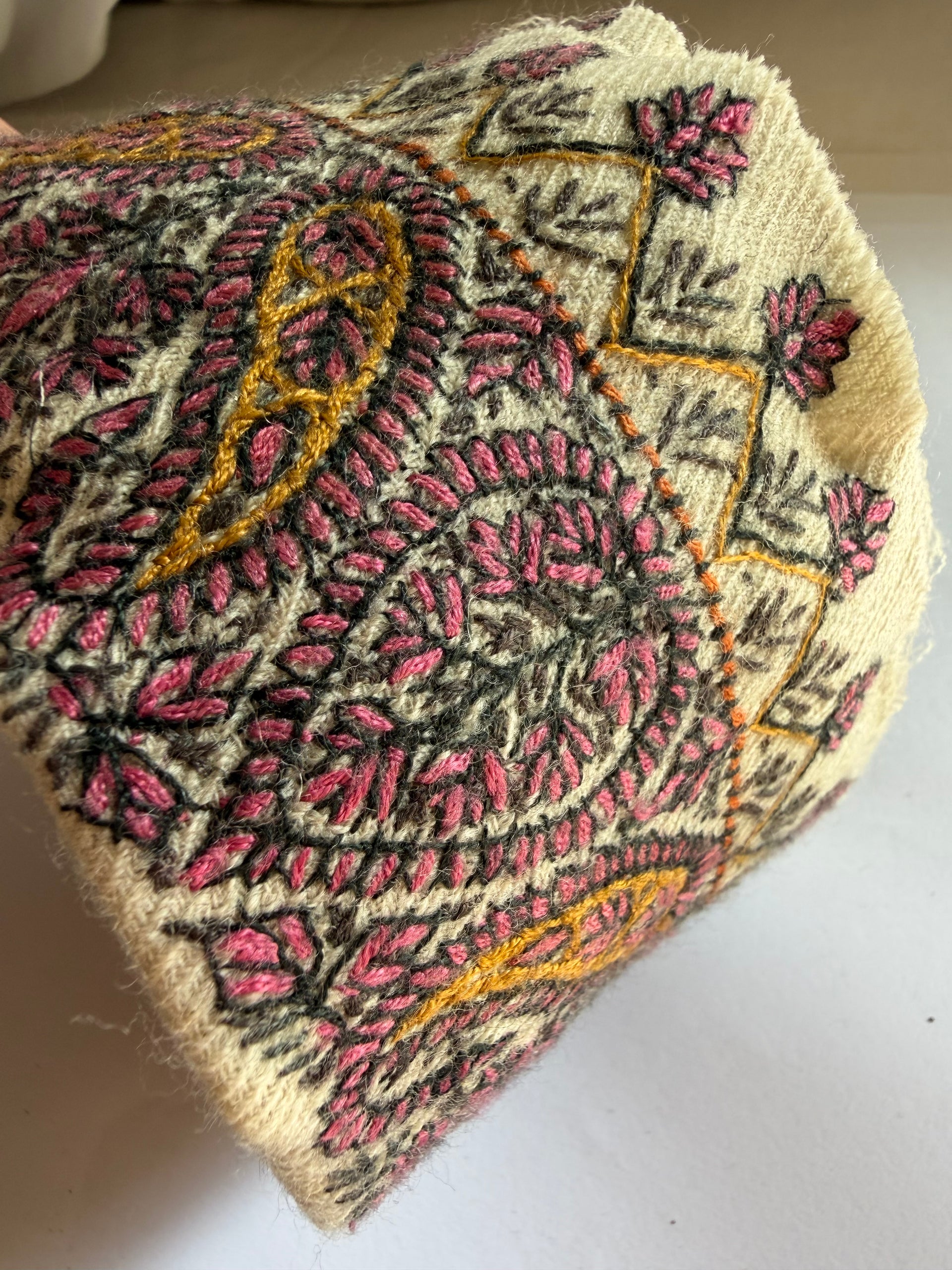 A close-up of a knitted or crocheted textile piece, likely a hat or bag, featuring a cream base with pink, black, and mustard yellow embroidered paisley and floral patterns.