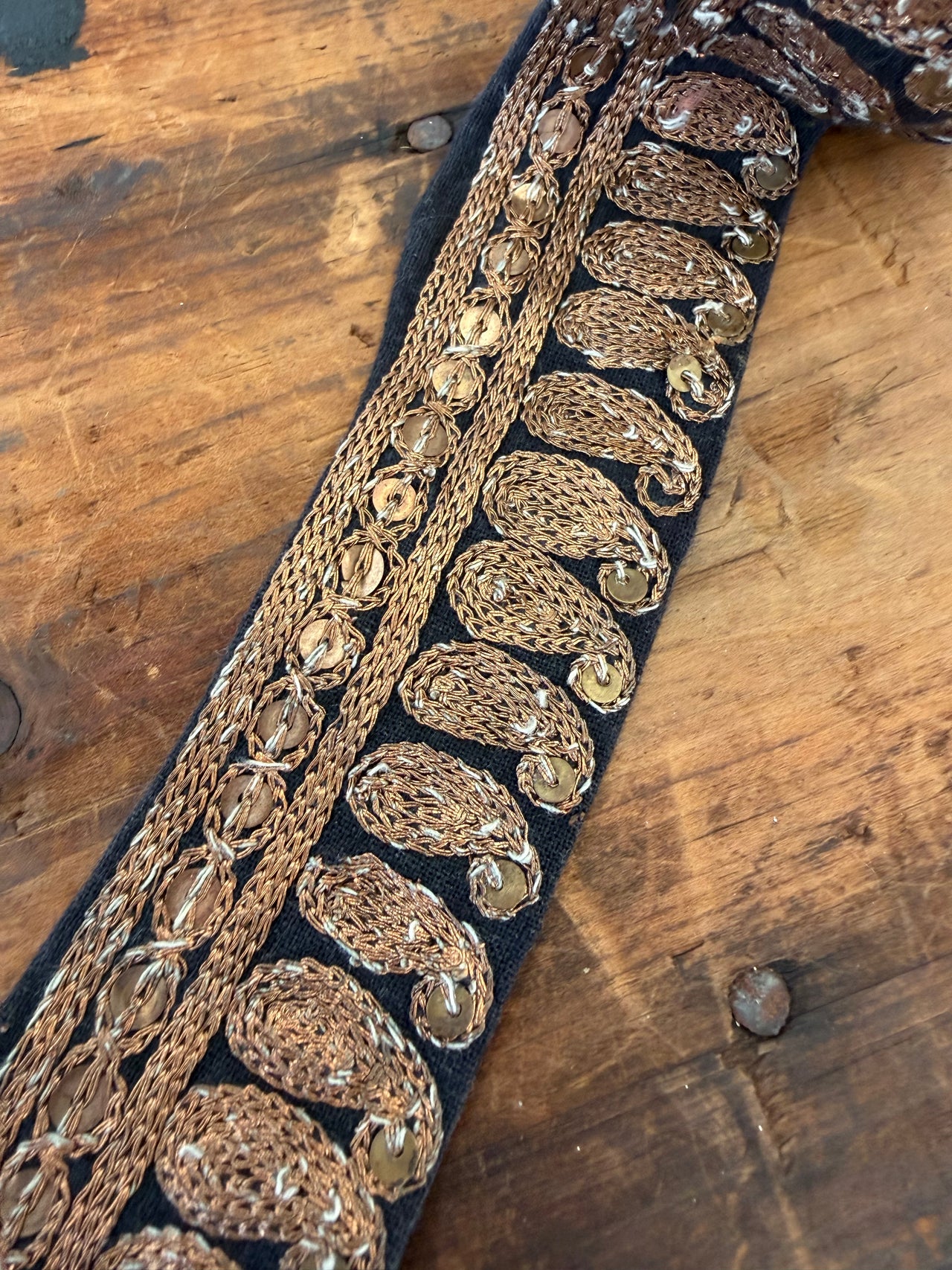 A close-up of an ornate embroidered trim featuring metallic gold paisley and braided patterns on a black fabric base, laid diagonally on a rustic wooden surface.