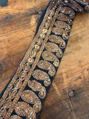 A close-up of an ornate embroidered trim featuring metallic gold paisley and braided patterns on a black fabric base, laid diagonally on a rustic wooden surface.
