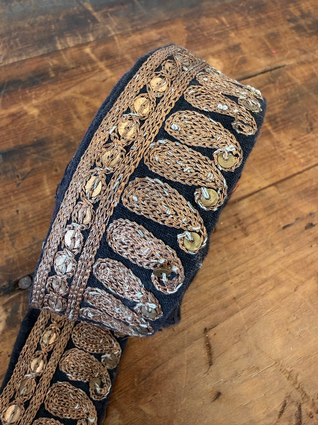 A close-up of a black fabric trim or belt featuring ornate metallic embroidery in gold, bronze, and silver, laid diagonally on a rustic wooden surface.