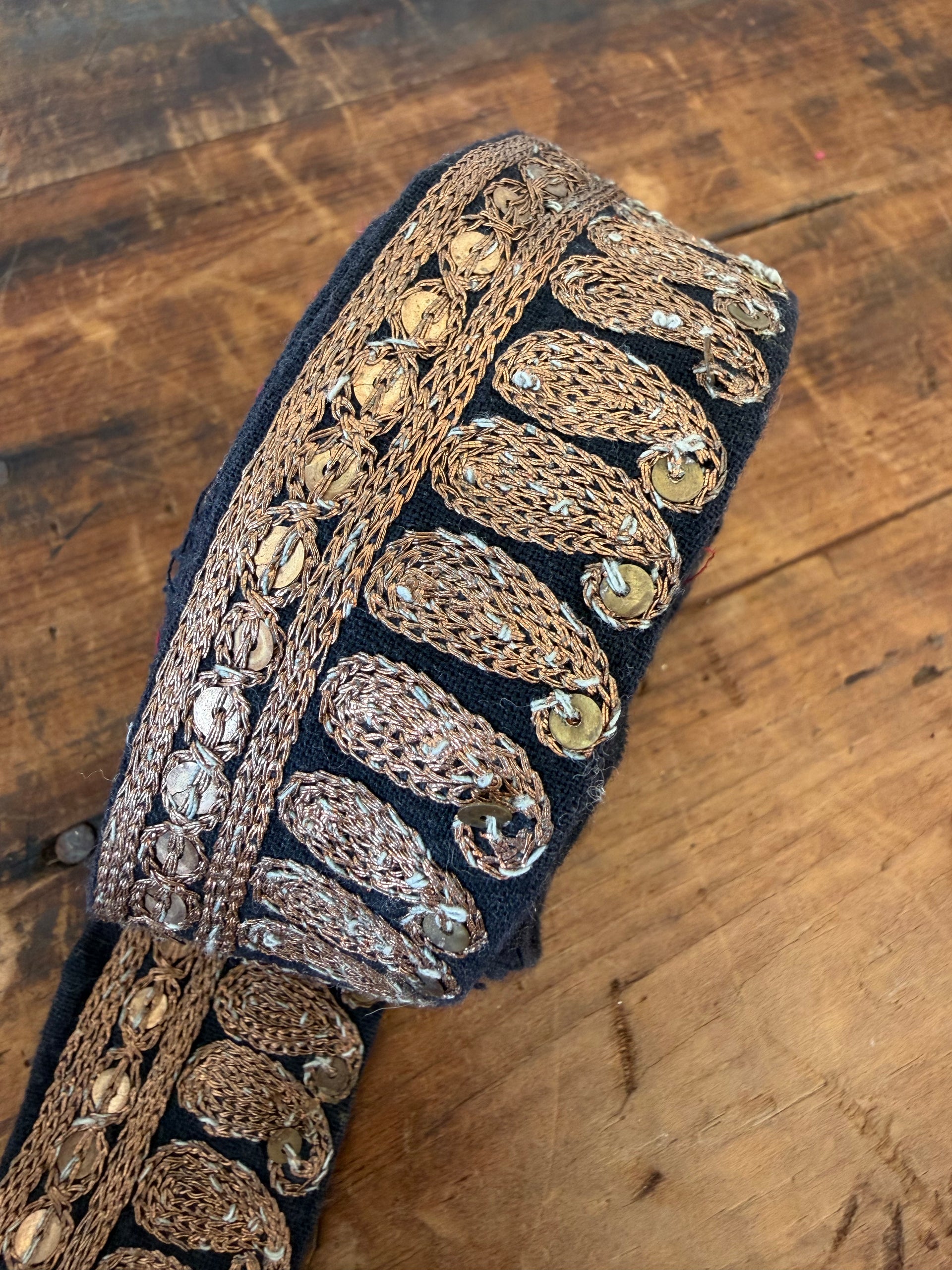 A close-up of a black fabric trim or belt featuring ornate metallic embroidery in gold, bronze, and silver, laid diagonally on a rustic wooden surface.