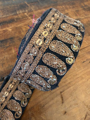 A close-up of an ornate embroidered trim or applique featuring metallic gold and bronze woven patterns with small reflective embellishments on a dark fabric base, laid on a wooden surface.