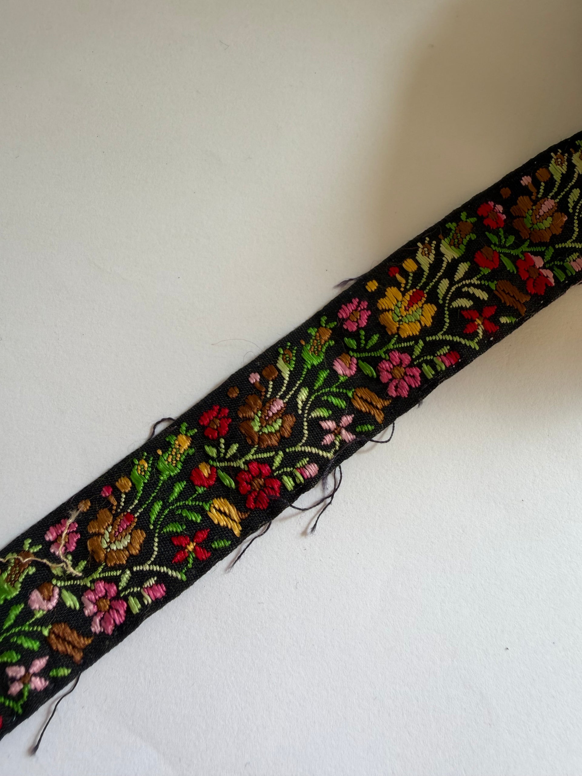 A close-up of a black embroidered trim featuring colorful floral patterns in red, pink, green, yellow, and brown, laid diagonally on a plain off-white background.