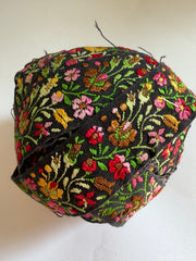 A round fabric headband or hair wrap featuring an intricate multicolor floral embroidery pattern on a black background, photographed against a plain off-white backdrop.