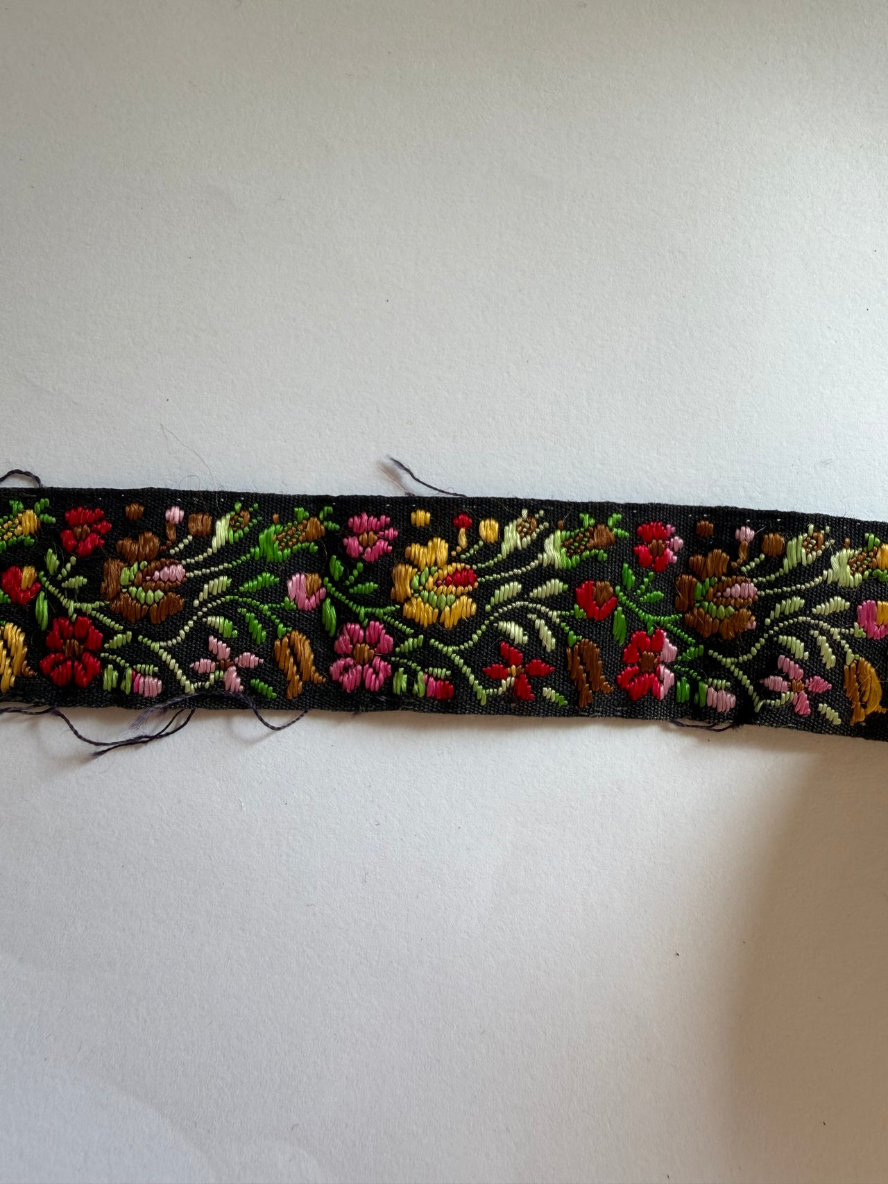 A vertical strip of black embroidered fabric trim featuring colorful floral patterns in red, pink, yellow, green, and brown, shown against a plain off-white background.