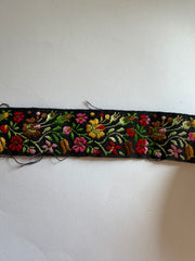A vertical strip of black embroidered fabric trim featuring colorful floral patterns in red, pink, yellow, green, and brown, shown against a plain off-white background.