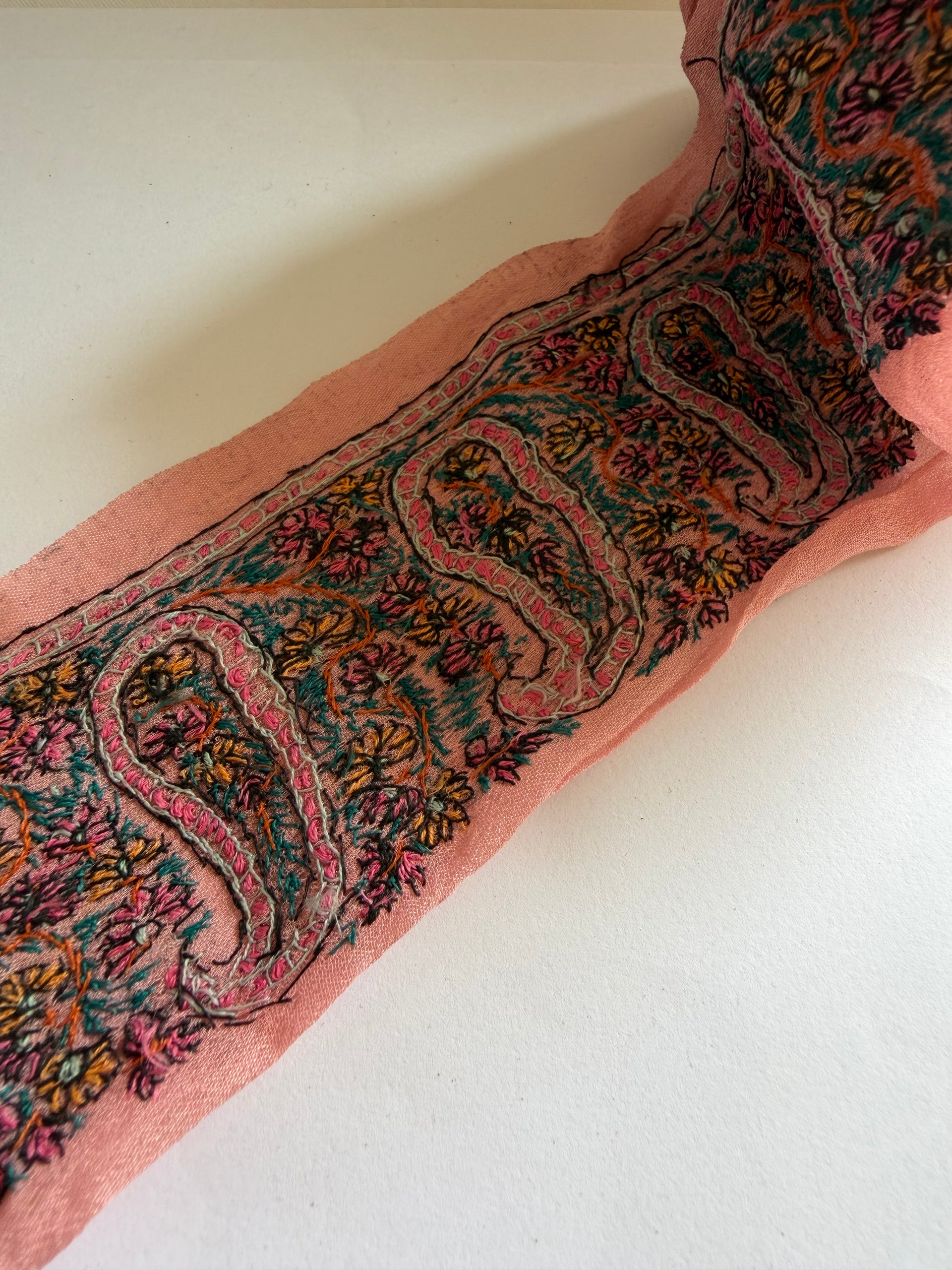 A close-up of a pink embroidered fabric or scarf with a detailed paisley and floral pattern in teal, gold, and purple, laid flat against a plain off-white background.