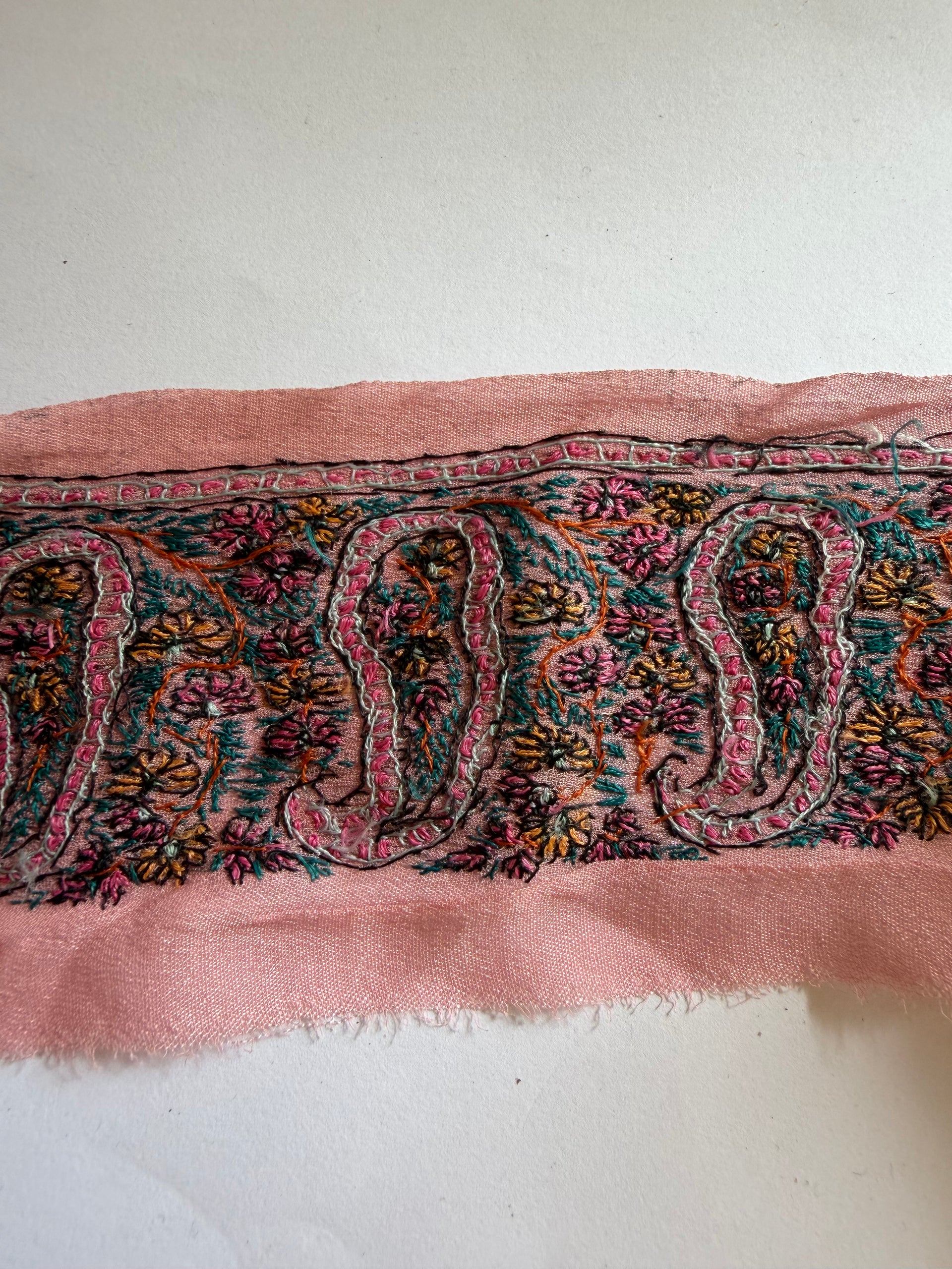 A close-up of a pink woven textile with a multicolor paisley and floral pattern, laid against a plain off-white background.