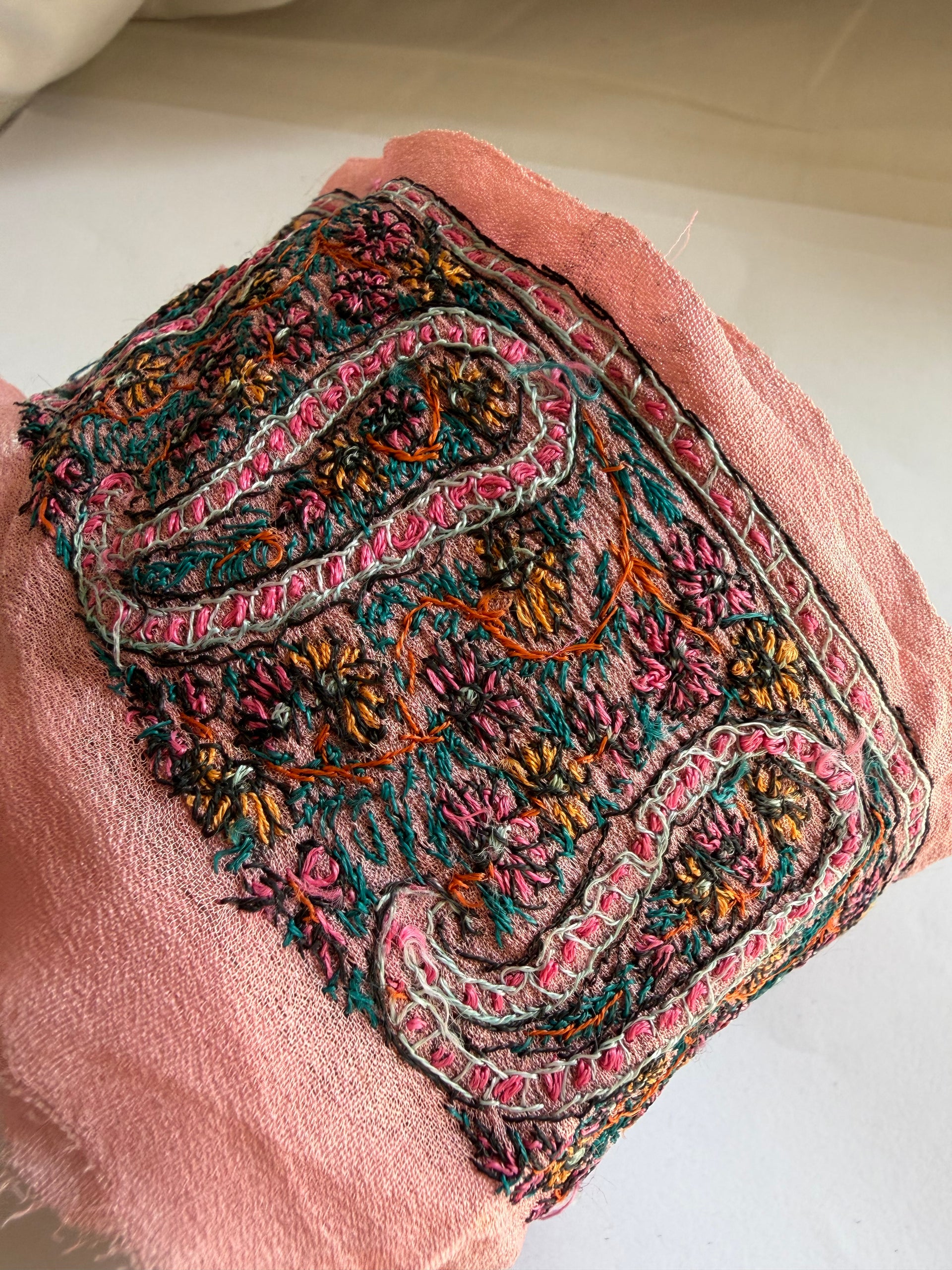 A close-up of a dusty pink fabric accessory, likely a scarf or shawl, featuring an intricate, hand-embroidered paisley and floral border in pink, teal, orange, and gold.