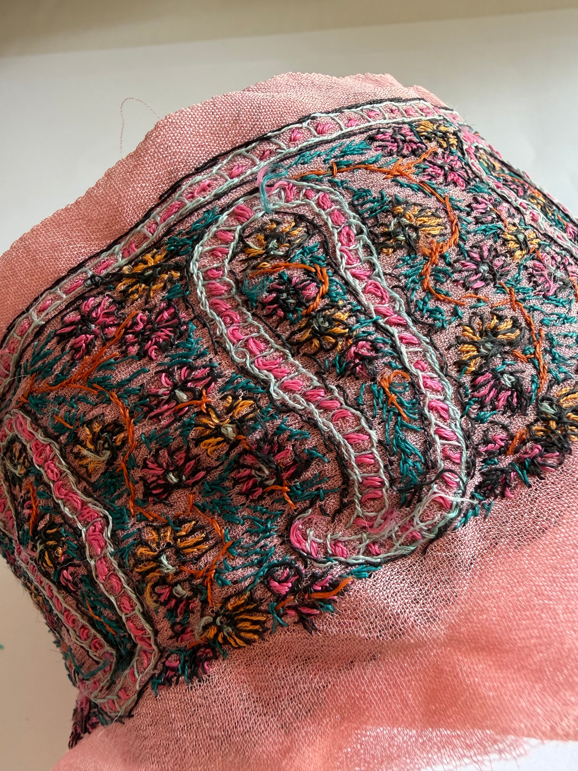 A close-up of a pink sheer mesh fabric with intricate multicolor embroidery, featuring floral and paisley-like patterns in pink, teal, orange, yellow, and black.