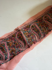 A close-up of a pink fabric trim or scarf featuring an intricate, multicolored paisley and floral pattern with black outlines, laid diagonally on a plain off-white background.