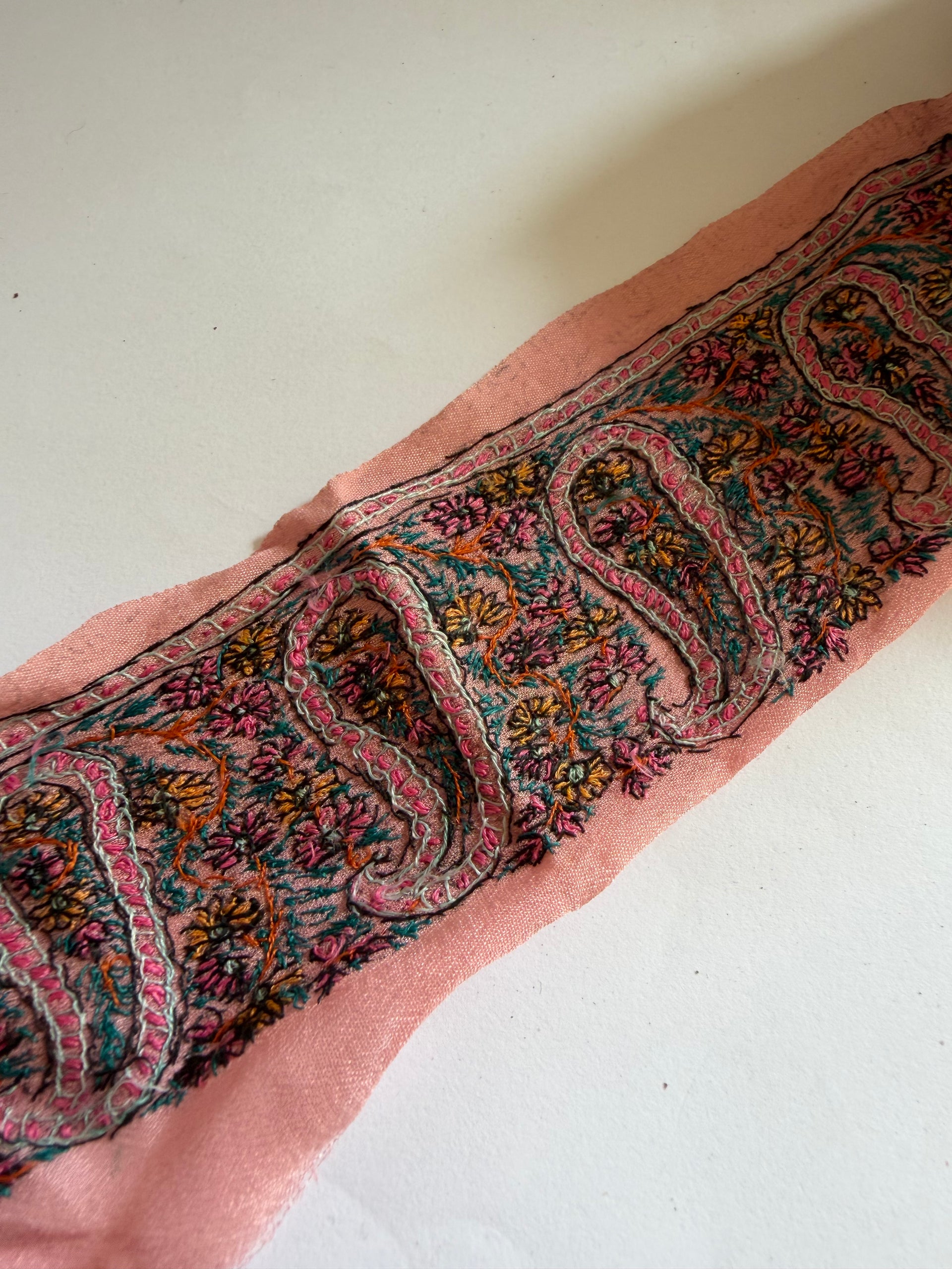 A close-up of a pink fabric trim or scarf featuring an intricate, multicolored paisley and floral pattern with black outlines, laid diagonally on a plain off-white background.