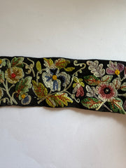 A close-up of a black embroidered trim or ribbon featuring multicolored floral motifs in gold, green, pink, blue, and red on a plain light background.