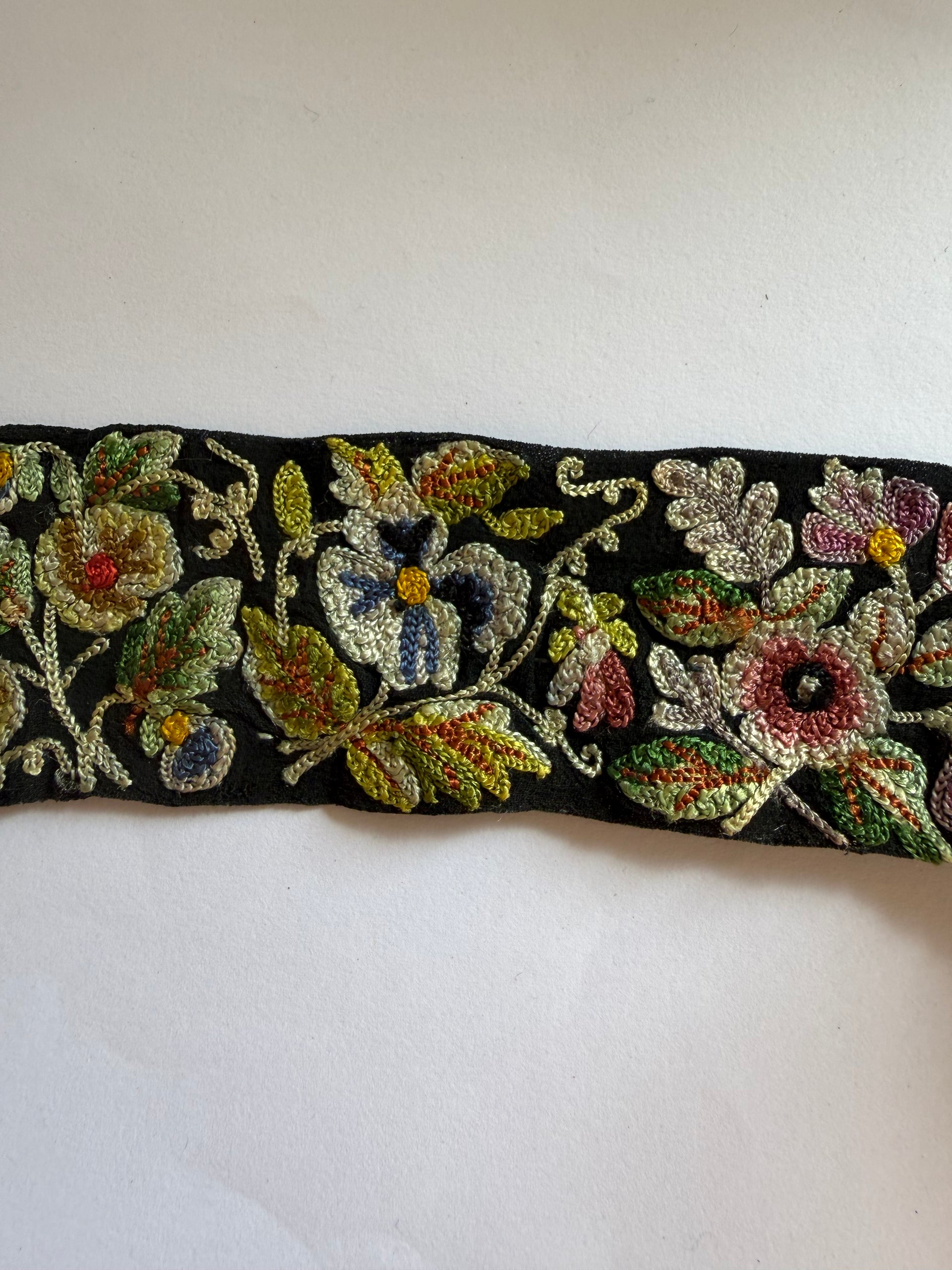 A close-up of a black embroidered trim or ribbon featuring multicolored floral motifs in gold, green, pink, blue, and red on a plain light background.