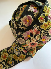 A close-up of a black fabric trim or ribbon featuring dense, colorful floral and leaf embroidery in pink, green, yellow, and beige tones, laid out on a plain off-white background.