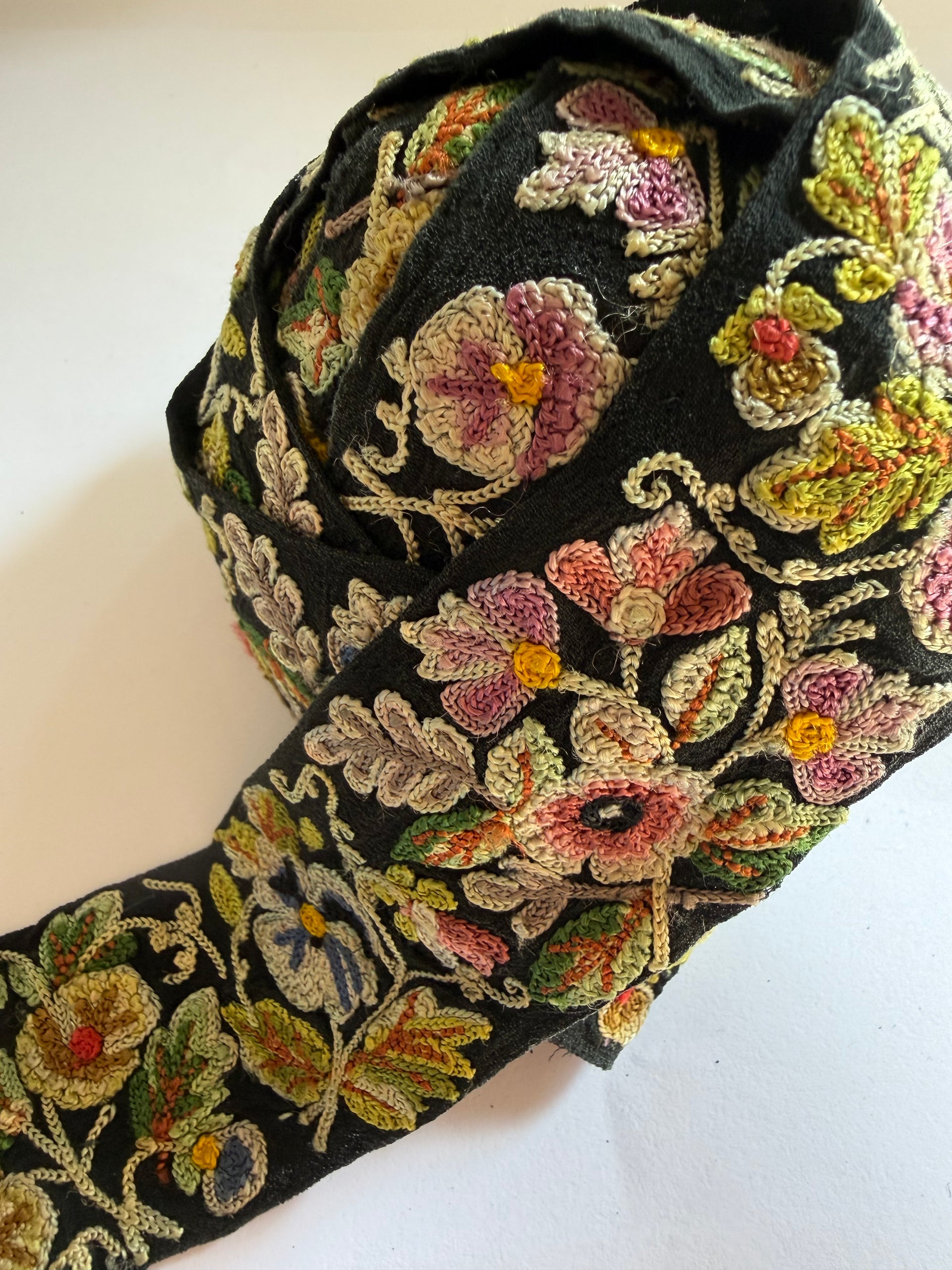 A close-up of a black fabric trim or ribbon featuring dense, colorful floral and leaf embroidery in pink, green, yellow, and beige tones, laid out on a plain off-white background.