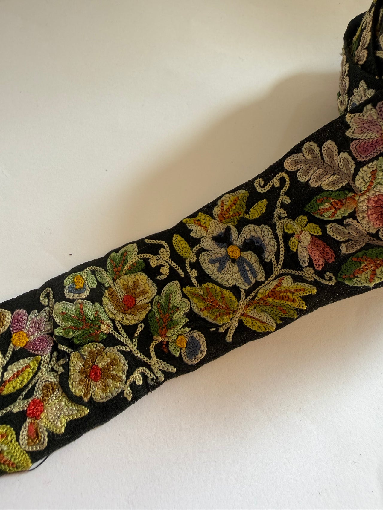 A close-up of a black embroidered fabric trim featuring intricate floral and leaf patterns in gold, green, pink, and blue thread, laid diagonally on a white background.