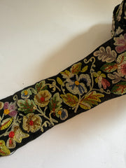 A close-up of a black embroidered fabric trim featuring intricate floral and leaf patterns in gold, green, pink, and blue thread, laid diagonally on a white background.
