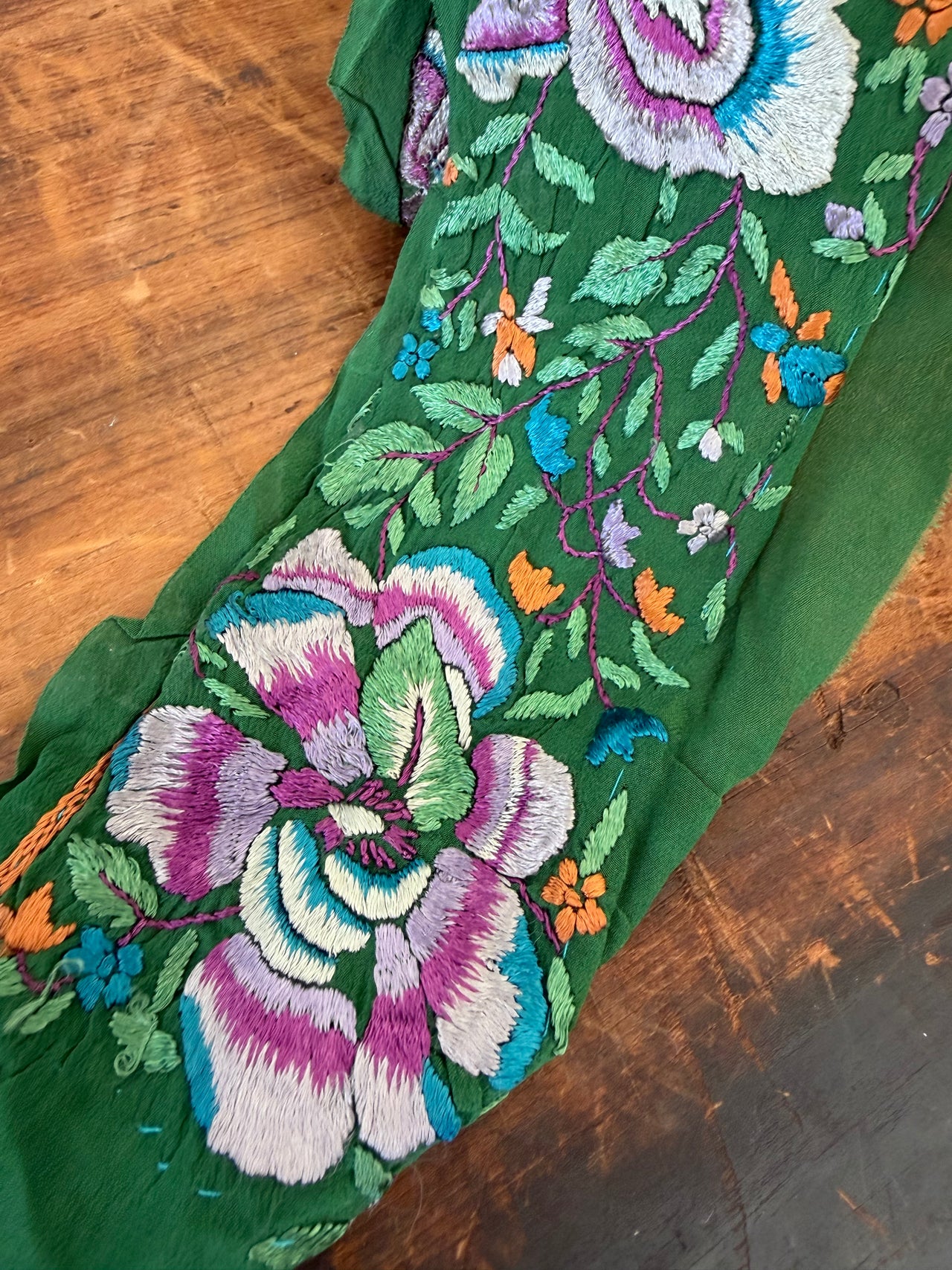 A close-up of a green embroidered fabric, likely a sleeve or garment panel, featuring large floral motifs in purple, white, blue, and orange on a rustic wooden background.