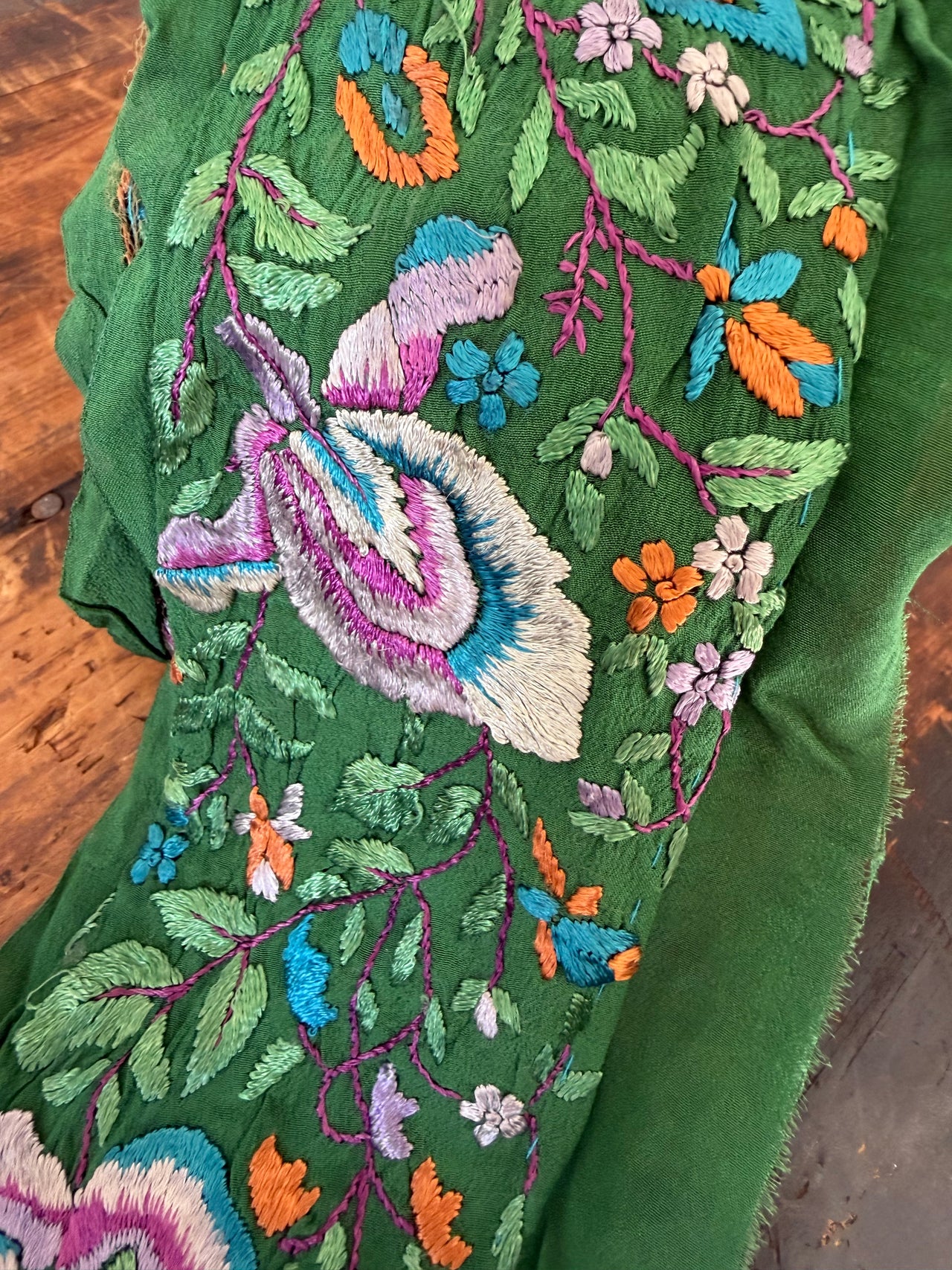 A close-up of a green embroidered textile featuring a large central floral motif in white, purple, and blue, surrounded by smaller flowers and leaves in orange, blue, pink, and green, laid over a brown wooden surface.