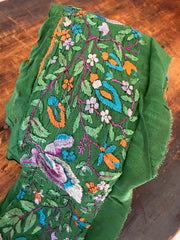 A folded green fabric panel featuring intricate multicolor embroidery with a butterfly, floral motifs, and paisley designs in purple, blue, orange, and white, laid on a dark wooden surface.