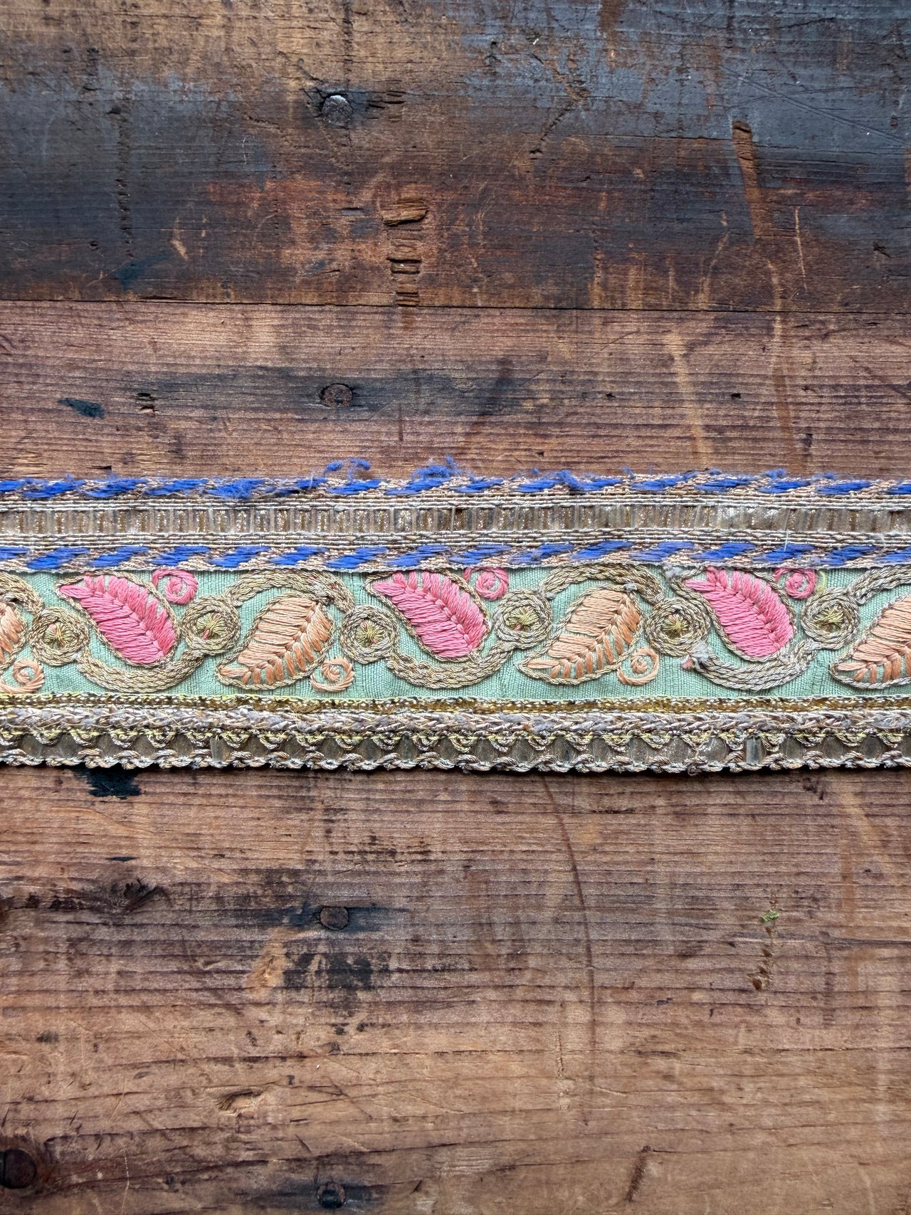 A decorative embroidered trim band laid horizontally across a rustic dark brown wooden plank background. The trim features pink and beige floral/leaf motifs on a mint green base, with blue and gold embroidered details along the top and bottom edges.