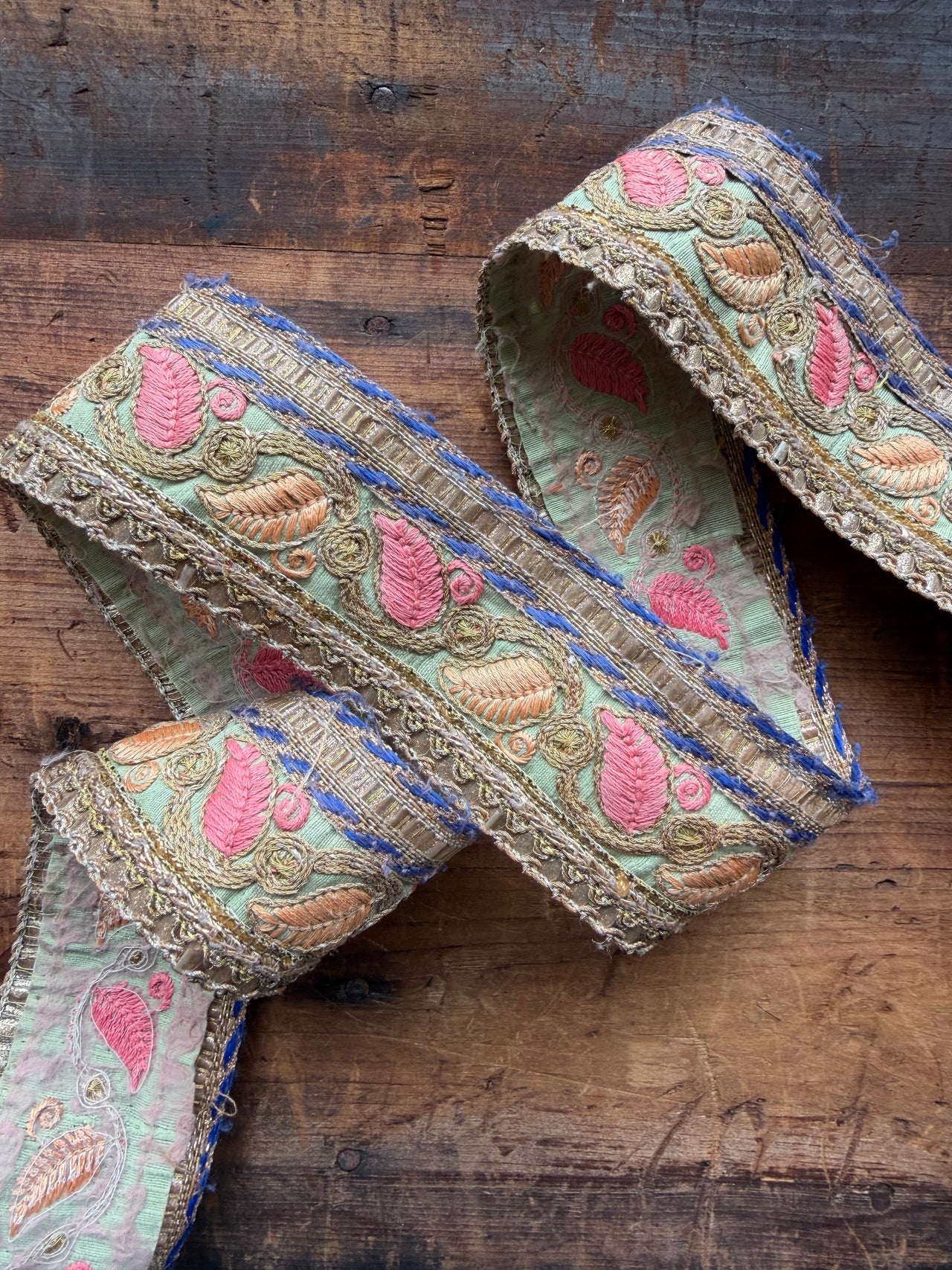 A decorative embroidered fabric trim ribbon with pink and gold paisley motifs on a sage green background, laid out on a rustic dark wood surface.