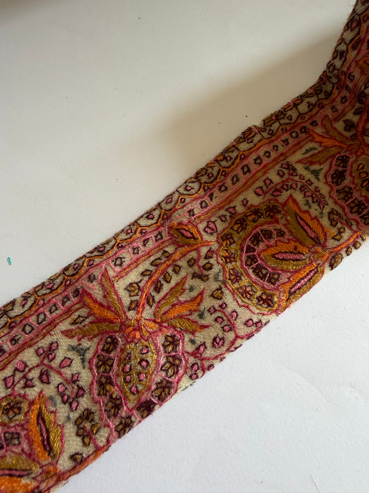 A close-up of a decorative embroidered fabric trim or applique featuring a detailed paisley and floral pattern in pink, orange, brown, and gold on a light beige background, laid diagonally on a white surface.
