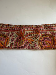 A close-up of a vintage patterned textile or scarf with intricate paisley and floral embroidery in pink, orange, brown, and gold on a cream background, laid flat against a white surface.