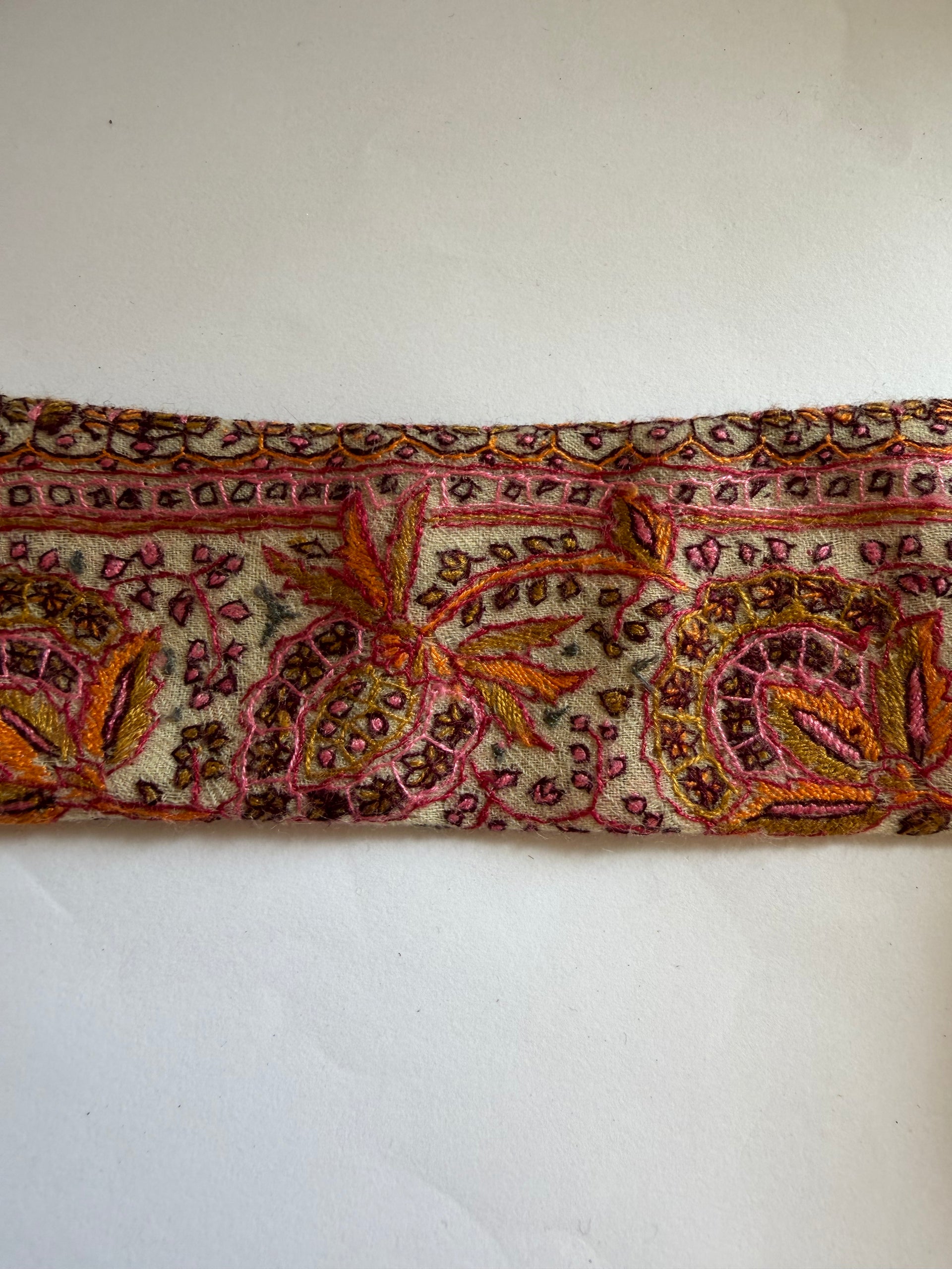 A close-up of a vintage patterned textile or scarf with intricate paisley and floral embroidery in pink, orange, brown, and gold on a cream background, laid flat against a white surface.