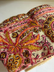 Close-up of an embroidered fabric accessory, likely a pouch or sleeve, featuring intricate floral and geometric patterns in pink, gold, brown, and orange on a cream-colored base.