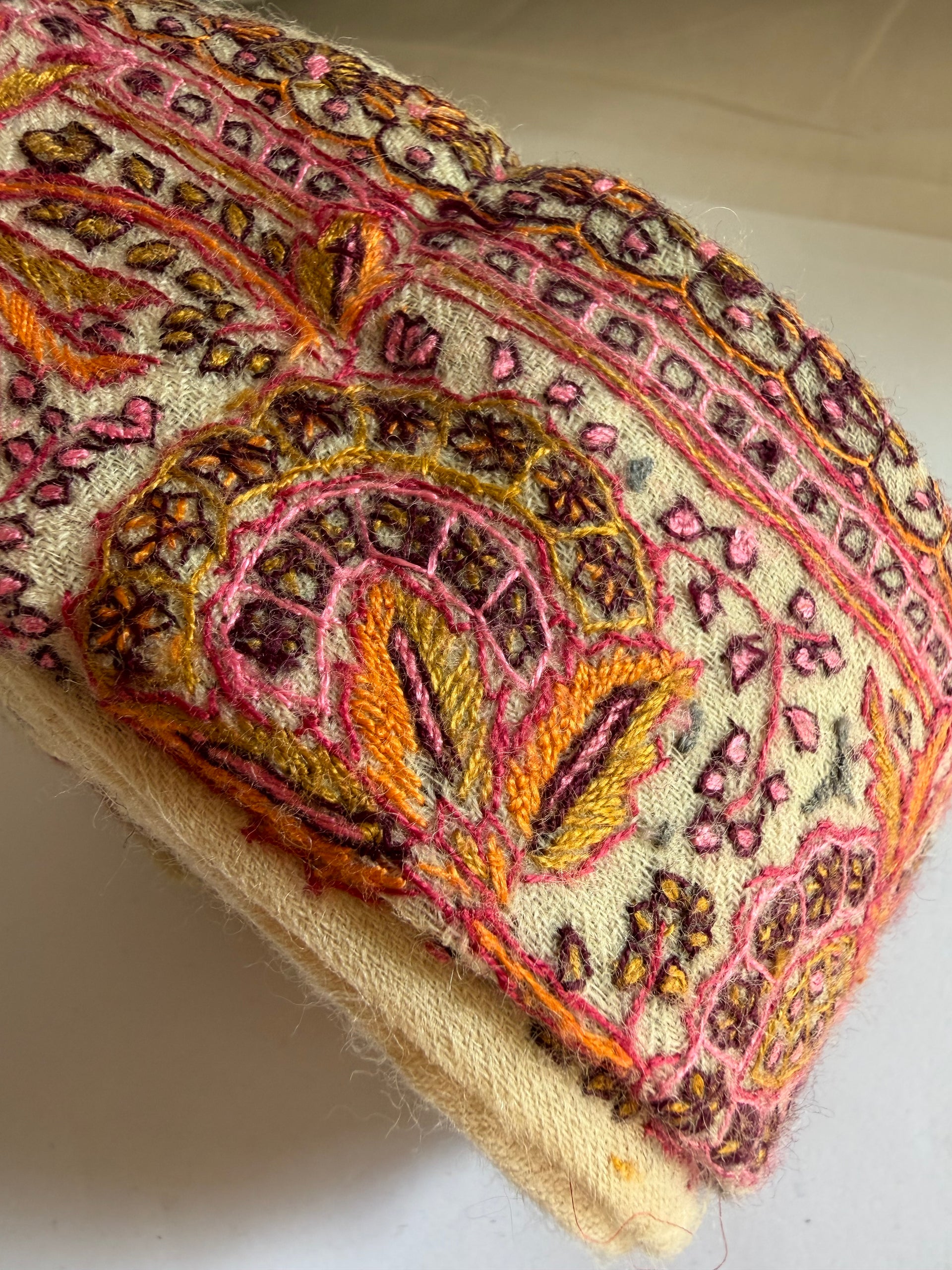 A close-up of a folded cream-colored fabric item, likely a scarf, shawl, or bandana, featuring intricate hand-embroidered paisley and floral patterns in pink, orange, yellow, and brown.