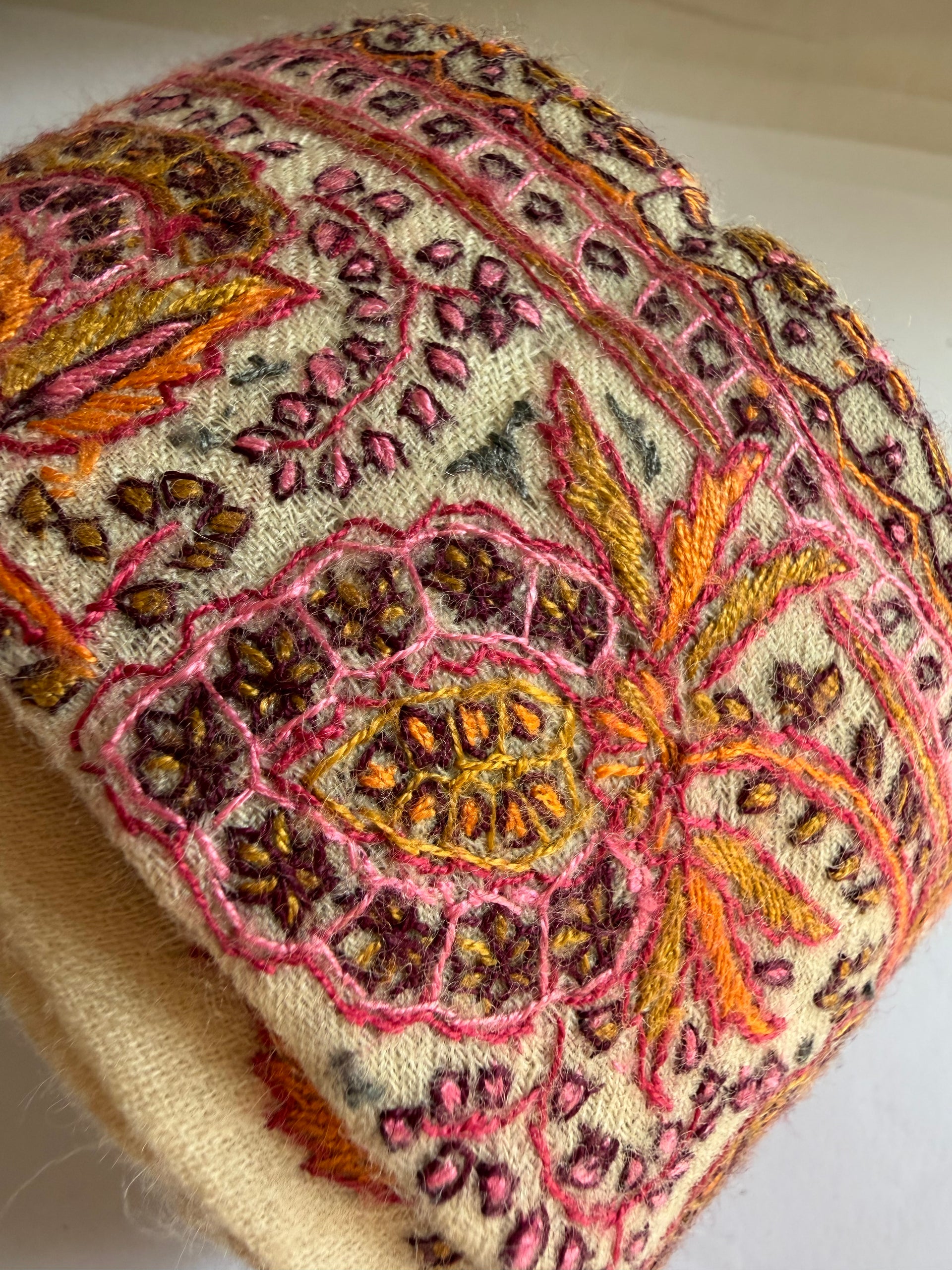 A close-up of a beige knit fabric item, likely a hat or scarf, heavily decorated with intricate hand-embroidered and stitched floral patterns in pink, orange, yellow, and brown.
