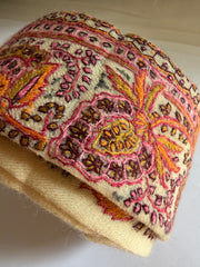 A close-up of a rolled piece of cream-colored fabric with intricate hand-embroidered or woven floral and paisley patterns in pink, orange, yellow, and brown.