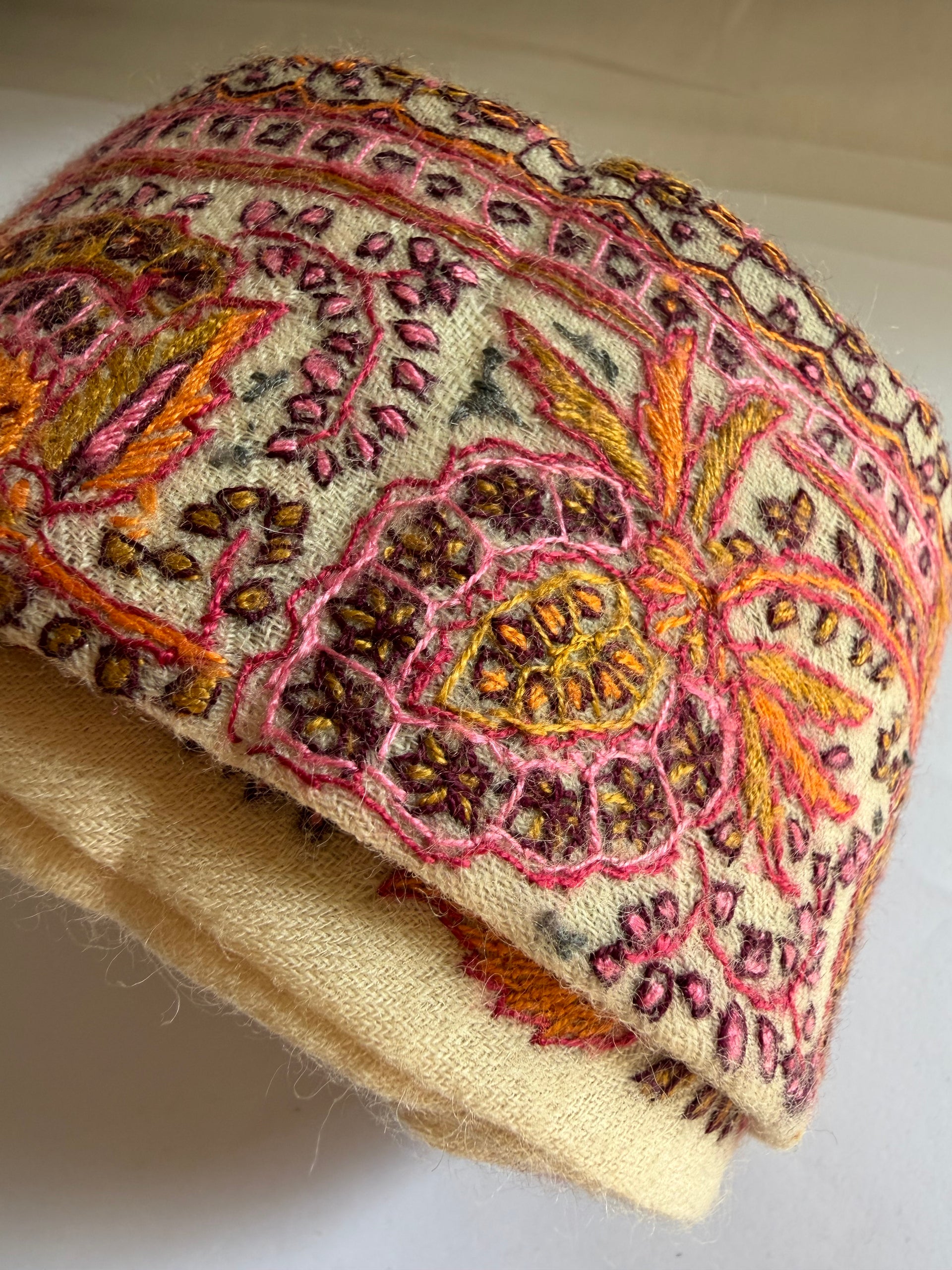 A close-up of a rolled piece of cream-colored fabric with intricate hand-embroidered or woven floral and paisley patterns in pink, orange, yellow, and brown.