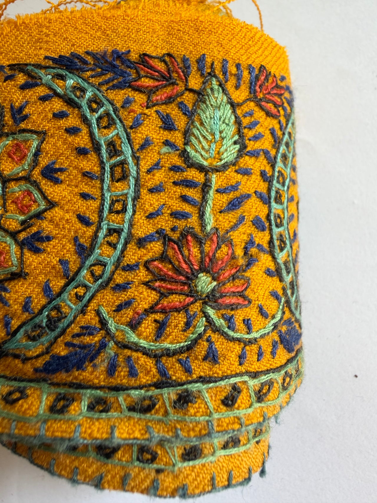 A close-up of a rolled piece of mustard yellow fabric with detailed multicolor embroidery featuring floral motifs in red, orange, and green, and paisley patterns in blue and green.