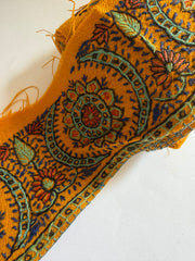 A close-up of a woven textile with a mustard yellow base and intricate multicolor floral and paisley patterns in blue, green, orange, and red, laid out on a white background.