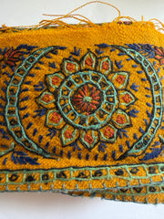 A close-up of a mustard yellow embroidered textile featuring a detailed central floral mandala pattern in teal, navy blue, and rust orange, with frayed edges on the left.