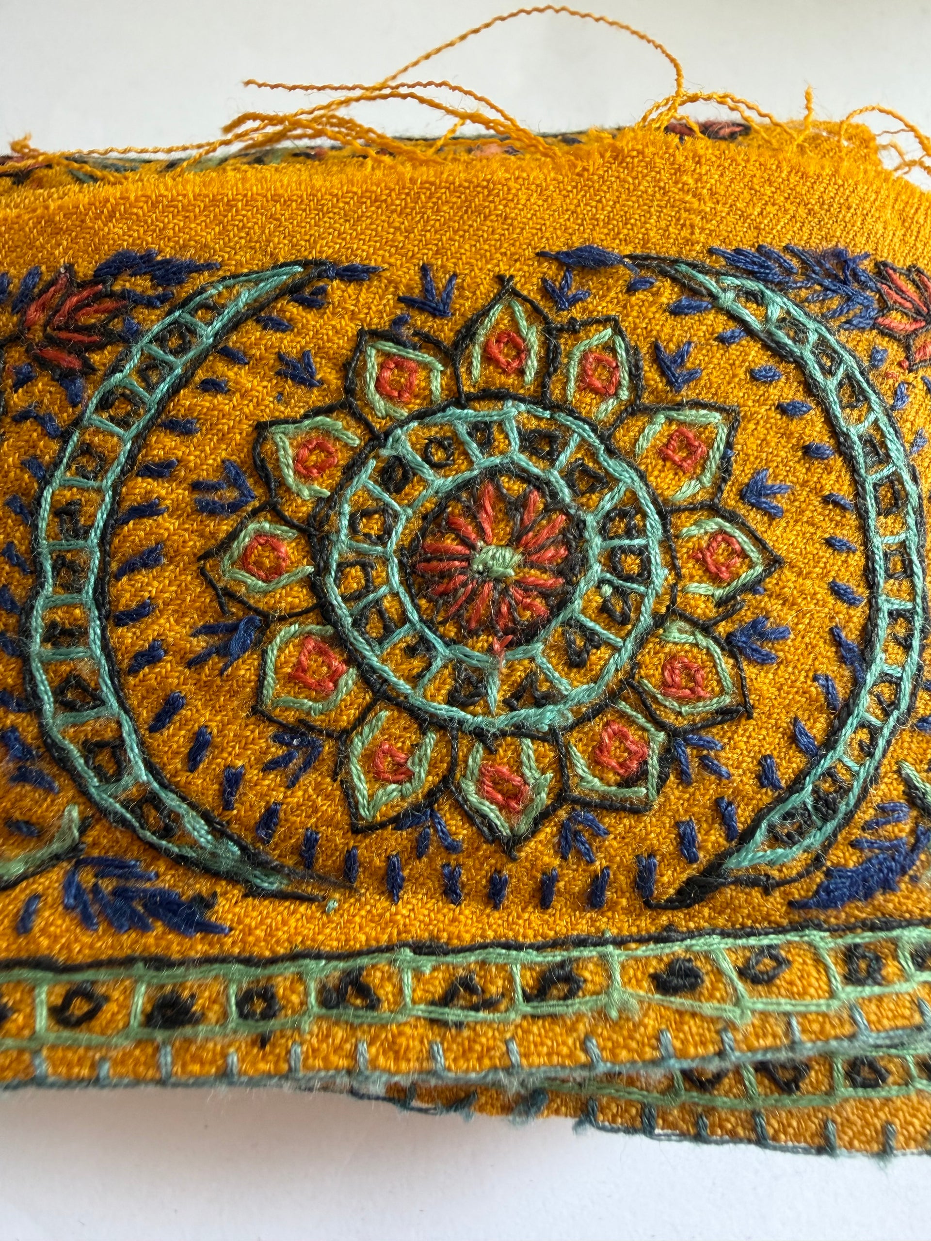 A close-up of a mustard yellow embroidered textile featuring a detailed central floral mandala pattern in teal, navy blue, and rust orange, with frayed edges on the left.