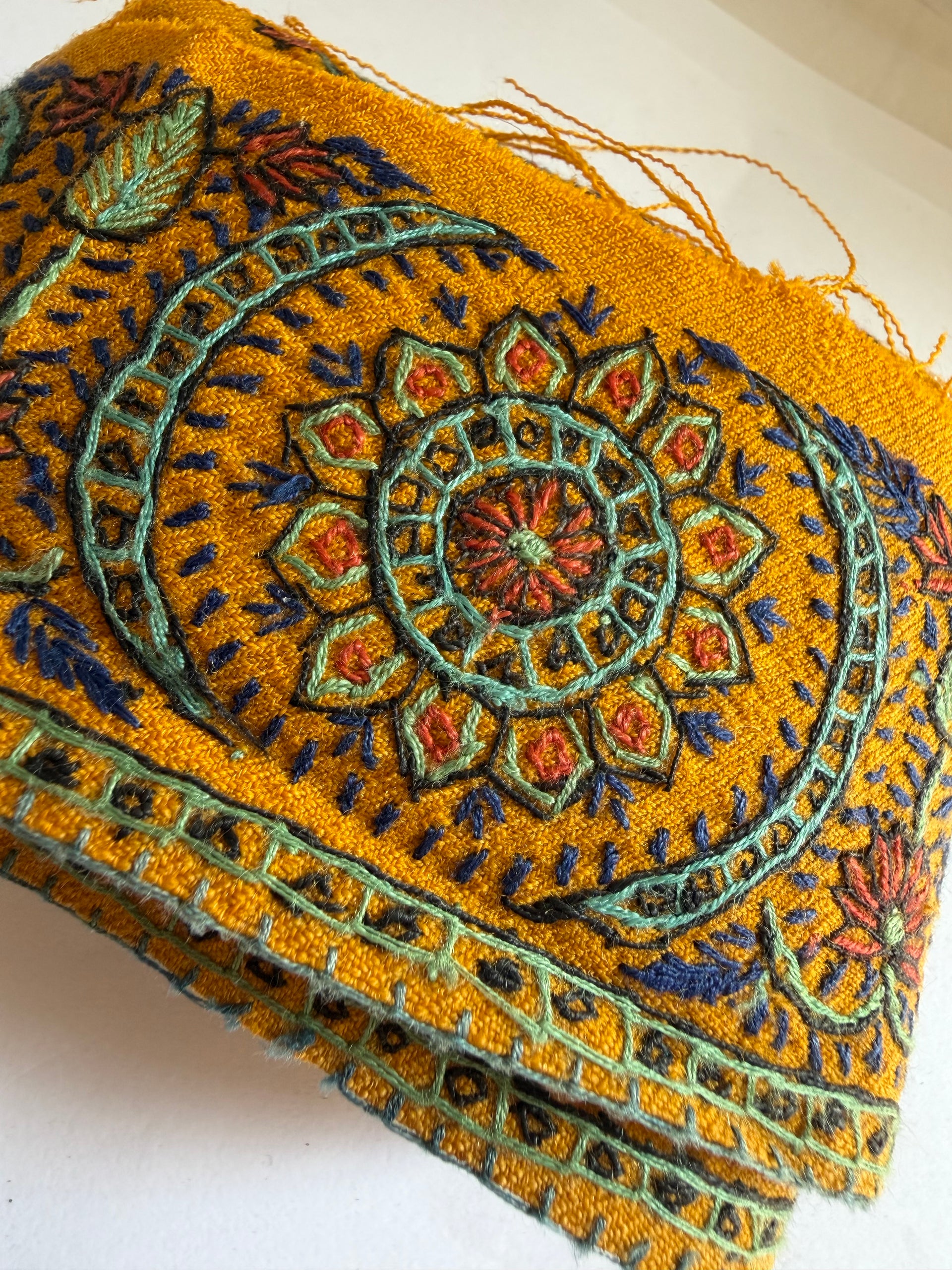 A close-up of a folded mustard yellow embroidered textile, likely a scarf, shawl, or decorative fabric, featuring intricate mandala and floral patterns in teal, blue, orange, and red.