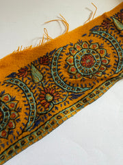 A close-up of a mustard yellow woven textile with an ornate embroidered or printed floral and geometric border pattern in blue, green, red, and orange, shown on a plain white background.