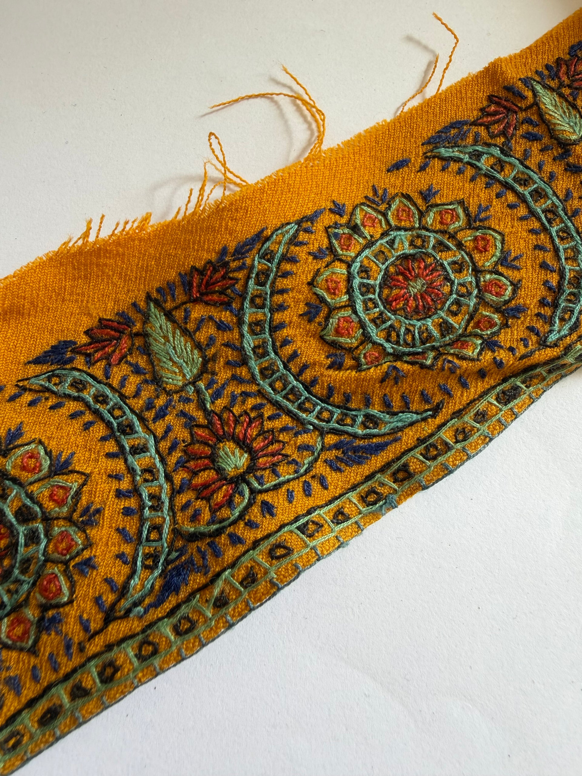 A close-up of a mustard yellow woven textile with an ornate embroidered or printed floral and geometric border pattern in blue, green, red, and orange, shown on a plain white background.