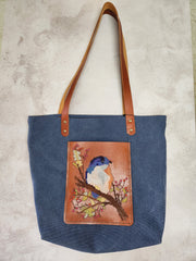 Cross-stitch tote bag with a bird design, blue fabric and tan leather handles